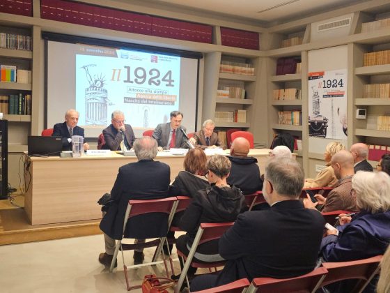 gruppo di persone riprese di spalle che partecipano un convegno, sullo sfondo i relatori dietro la scrivania