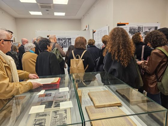 gruppo di persone riprese di spalle che partecipano a un'inaugurazione di una mostra, in primo piano teche espositive con materiale librario