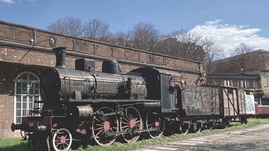 Un treno a vapore sui binari, in primo piano la locomotiva di colore scuro, in secondo piano un edificio con tetto basso, sullo sfondo il cielo azzurro.